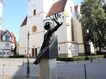 Herford-Fürstenaudenkmal-Pro Herford GmbH.JPG Bronzeskulptur eines Mannes mit erhobenem Arm, vor einer historischen Kirche in einer ruhigen Straße.