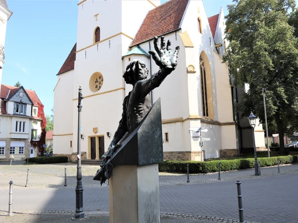 Herford-Fürstenaudenkmal-Pro Herford GmbH.JPG Bronzeskulptur eines Mannes mit erhobenem Arm, vor einer historischen Kirche in einer ruhigen Straße.