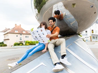 Ein Paar sitzt vor einer großen, spiegelnden Kugel in einer urbanen Umgebung und hält eine Karte.