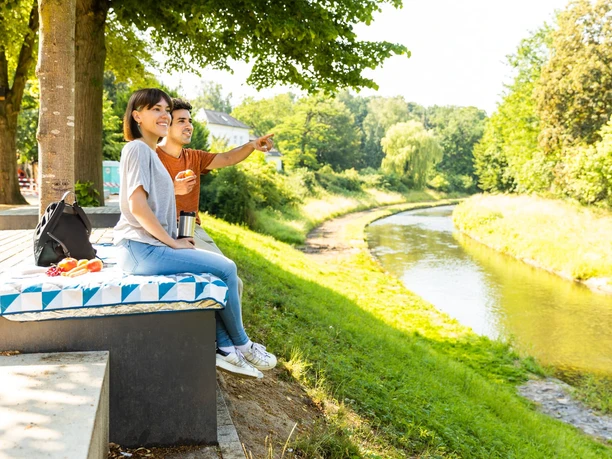 herford-promenade-teutoburger-wald-tourismus-h-tornow-023-jpg.jpg Zwei Personen sitzen auf einer Bank an einem Fluss, umgeben von grüner Natur und strahlendem Sonnenschein.