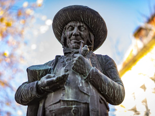Bronzestatue eines älteren Mannes mit Pfeife und Hut vor herbstlichem Hintergrund im Sonnenschein.