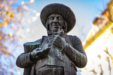 Herford_Herbst_Pro Herford GmbH.jpg Bronzestatue eines älteren Mannes mit Pfeife und Hut vor herbstlichem Hintergrund im Sonnenschein.