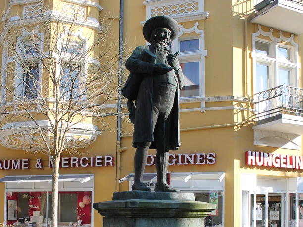 Statue eines Mannes mit Hut in traditioneller Kleidung vor einem gelben Gebäude in der Stadtmitte.