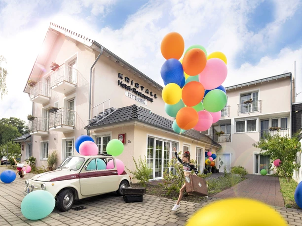 Kristallhotel FETTEHENNE in Leverkusen Ein modernes Hotelgebäude mit bunten Luftballons und einem Oldtimer, umgeben von klarer, blauer Himmel.