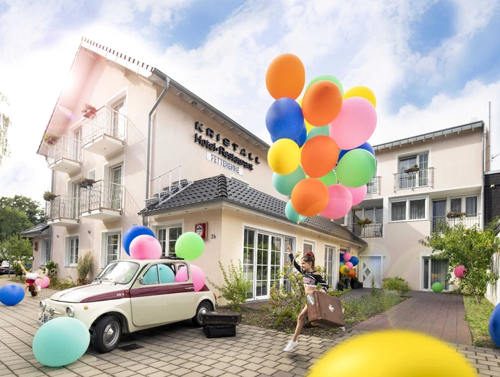 Kristallhotel FETTEHENNE in Leverkusen Ein modernes Hotelgebäude mit bunten Luftballons und einem Oldtimer, umgeben von klarer, blauer Himmel.