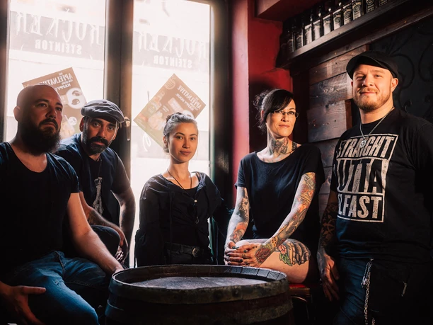 The Hoochigans Fünf tätowierte Personen posieren lächelnd in einem dunklen, stilvollen Raum mit Holzwänden und Fenstern.