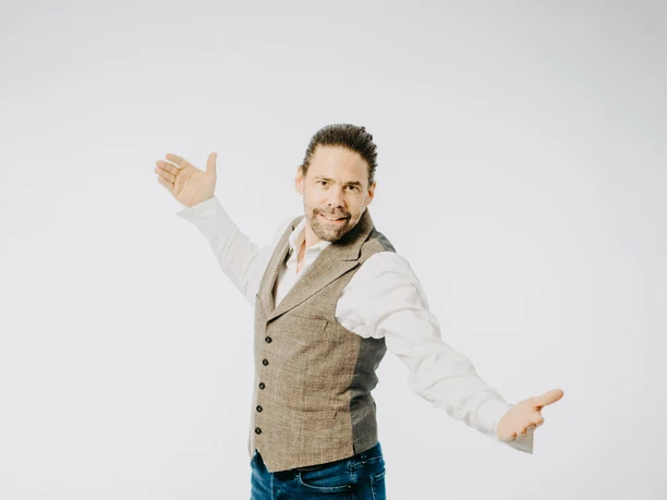 DJ Hendrik Treuse-31.jpg DJ Hendrik Treuse steht mit ausgebreiteten Armen lächelnd vor einem neutralen, hellen Hintergrund.
