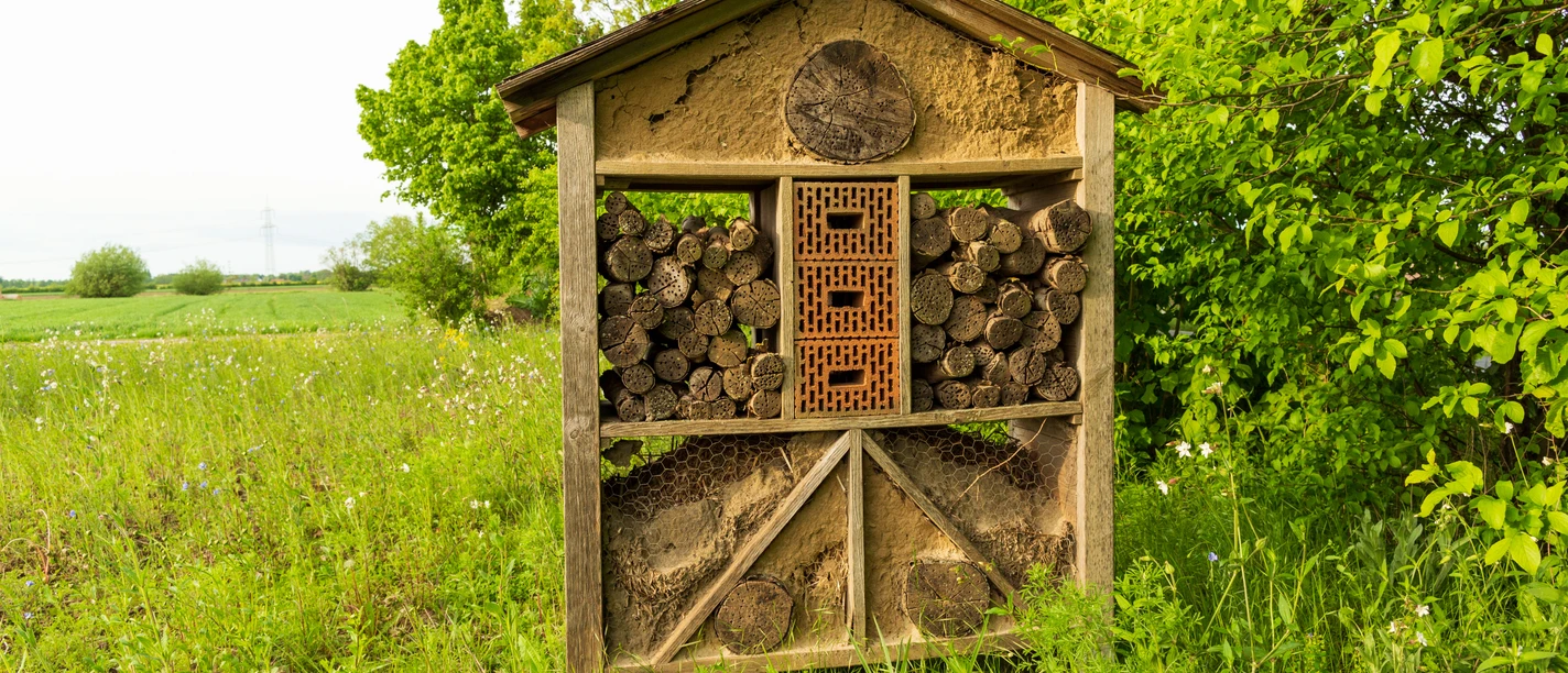 Ein Insektenhotel aus Holz und Lehm vor einem grünen Feld und Bäumen bei leicht bewölktem Himmel.