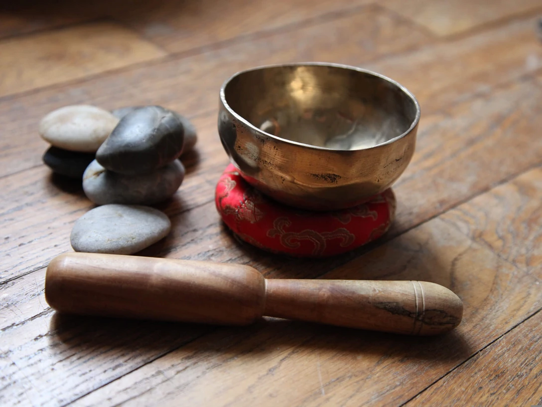 Klangreise.jpg Goldene Klangschale auf rotem Kissen mit Holzklöppel und glatten Steinen auf Holzuntergrund.