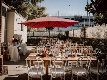 C5416C42C03C4D118E600BCB723BFCC3.jpg Holztische in Reihen mit rosaner Blumendeko auf der Terrasse mit SonnenschirmWooden tables in rows with pink floral decorations on the terrace with parasol