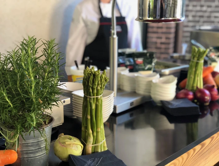 Markos Catering 1.jpeg Buffet Station mit Thymian, Spargel und weißen Tellern, dahinter steht eine Person mit schwarzer SchürzeBuffet station with thyme, asparagus and white plates, behind it stands a person with a black apron