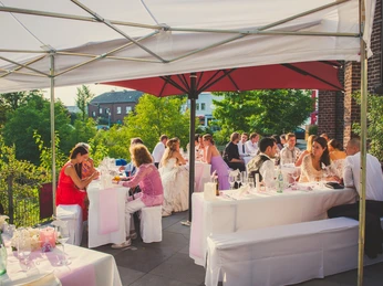 Eventkapelle - Hochzeit Susanne & Jojo (65) Kopie.jpg Hochzeitsgäste sitzen an weiß gedeckten Tischreihen auf der begrünten Außenterrasse der Eventkapelle unter SonnenschirmenWedding guests sit at rows of tables set in white on the green outdoor terrace of the event chapel under parasols