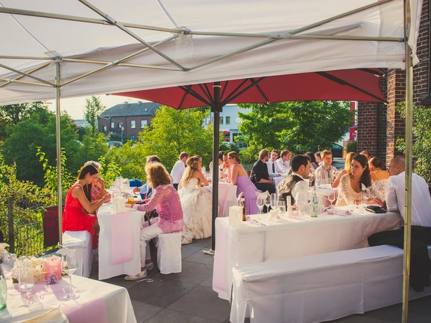Eventkapelle - Hochzeit Susanne & Jojo (65) Kopie.jpg Hochzeitsgäste sitzen an weiß gedeckten Tischreihen auf der begrünten Außenterrasse der Eventkapelle unter Sonnenschirmen