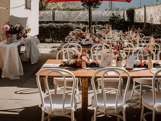 Hochzeit auf der Terasse.png Terrasse der Eventkapelle mit Holztischen, die mit Rosendeko für eine Hochzeit geschmückt sind