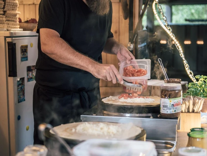 image 2.png Crepes Station: Ein schwarz gekleideter Mann mit langem dunkelbraunen Bart belegt vor sich liegende CrepesCrepes station: A man dressed in black with a long dark brown beard places crepes in front of him