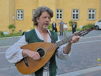 Musiker in historischer Kleidung spielt Laute vor gelber Gebäude-fassade mit gepflastertem Hof.