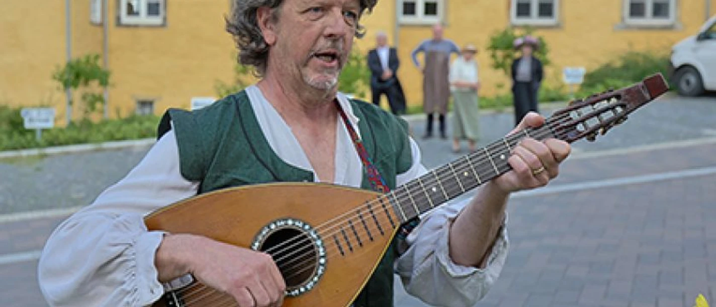 Musik Lübbecke Musiker in historischer Kleidung spielt Laute vor gelber Gebäude-fassade mit gepflastertem Hof.