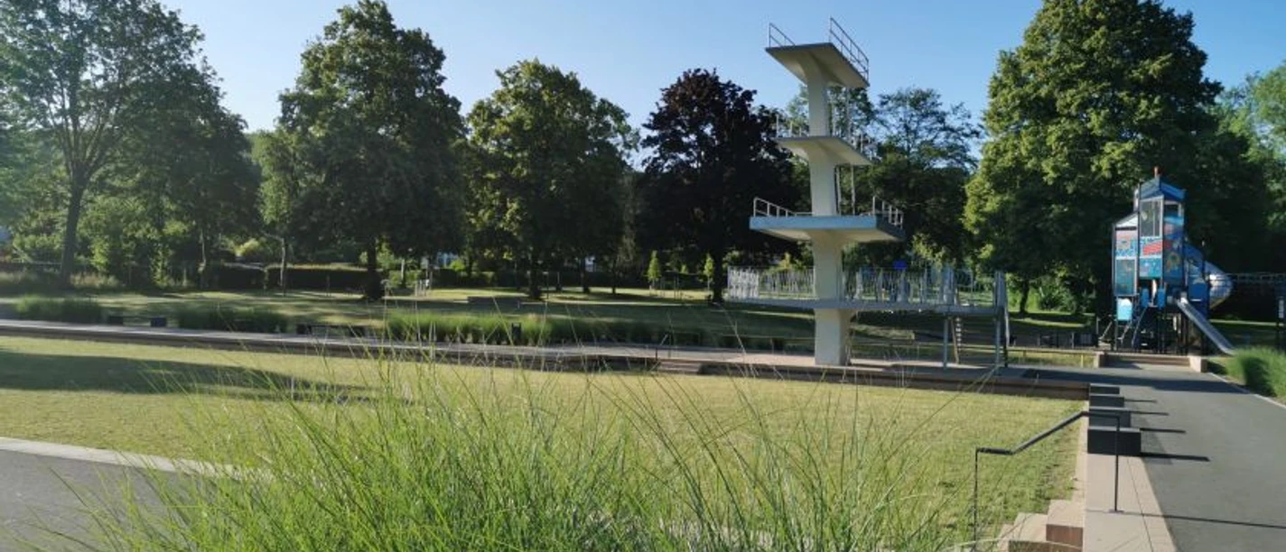 Grünes Freibad mit Sprungturm im Hintergrund, umgeben von schattenspendenden Bäumen und Rasenflächen.