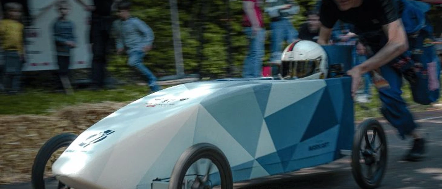 In einer blauen, spitz zulaufenden Seifenkiste auf vier Rädern, sitzt eine Person mit Motorradhelm. Sie wird von einer Person angeschoben.