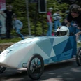 Seifenkistenrennen (c) Hannes Schmidt_Steve Edwards.jpg In einer blauen, spitz zulaufenden Seifenkiste auf vier Rädern, sitzt eine Person mit Motorradhelm. Sie wird von einer Person angeschoben.