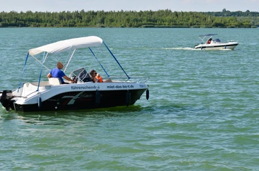 La photo montre deux bateaux en mouvement sur le lac de Störmthal, sur lesquels se trouvent plusieurs personnes. On remarque le temps de rêve qui se caractérise par une vue claire, un ciel sans nuages et un soleil radieux. Les bateaux portent les inscriptions "sans permis" et "miet-das-boot.de" et peuvent être loués au port de Störmthal.