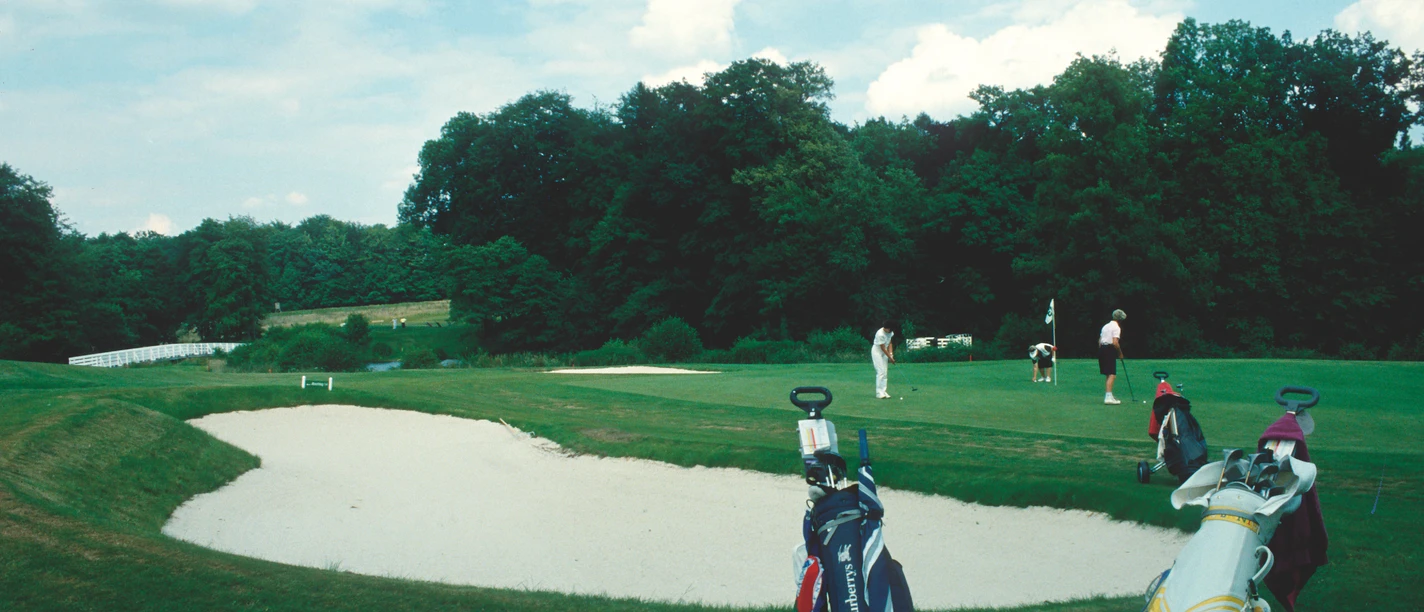 Golfer schlagen Bälle auf einem gepflegten Golfplatz mit Sandbunker, umgeben von Bäumen und blauen Himmel.