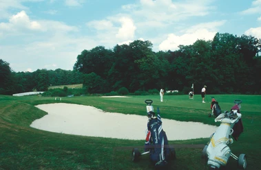 Senne-Golfclub_SHS_CC-BY-SA_Teutoburger_Wald_Stadt_Schloss_Holte-Stukenbrock Golfer schlagen Bälle auf einem gepflegten Golfplatz mit Sandbunker, umgeben von Bäumen und blauen Himmel.