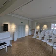 Ringelnatz Geburtshaus Saal Das Bild stellt einen Veranstaltungsaal mit einem am Eingang platzierten weißen Klavier und Stühle die nach diesem augerichtet sind, ab. In dem Raum hängen zudem einige Kunstwerke aus.The picture depicts an event hall with a white piano placed at the entrance and chairs aligned towards it. There are also some works of art hanging in the room.Na obrázku je zobrazen sál s bílým klavírem umístěným u vchodu a židlemi vyrovnanými směrem k němu. V místnosti visí také několik uměleckých děl.Obraz przedstawia salę eventową z białym fortepianem umieszczonym przy wejściu i krzesłami ustawionymi w jego kierunku. W pomieszczeniu wiszą również dzieła sztuki.De foto toont een evenementenhal met een witte piano bij de ingang en stoelen die ernaar toe zijn gericht. Er hangen ook enkele kunstwerken in de ruimte.L'immagine ritrae una sala per eventi con un pianoforte bianco all'ingresso e sedie allineate verso di esso. Nella sala sono appese anche alcune opere d'arte.