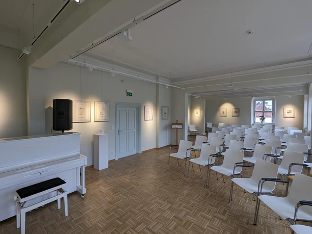 Ringelnatz Geburtshaus Saal Das Bild stellt einen Veranstaltungsaal mit einem am Eingang platzierten weißen Klavier und Stühle die nach diesem augerichtet sind, ab. In dem Raum hängen zudem einige Kunstwerke aus.