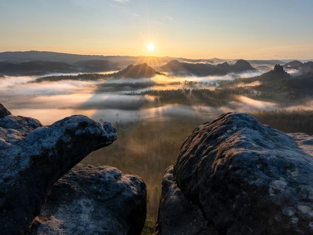 Sonnenaufgang über einer nebligen Landschaft mit bewaldeten Hügeln, im Vordergrund sind dunkle Felsen sichtbar.