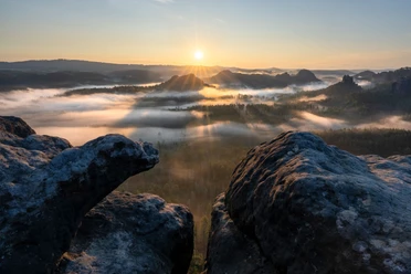 Sonnenaufgang über einer nebligen Landschaft mit bewaldeten Hügeln, im Vordergrund sind dunkle Felsen sichtbar.