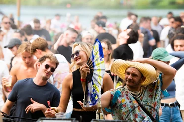 EAT PLAY LOVE Festival Ein fröhliches Festivalpublikum tanzt ausgelassen im Freien. Eine Person in einem farbenfrohen Outfit und großem Strohhut fächert mit einem Fächer Luft zu, während die umgebenden Menschen mit Sonnenbrillen im Sommerlook die Musik genießen. Im Hintergrund verschwimmen blauer Himmel und grüne Bäume.