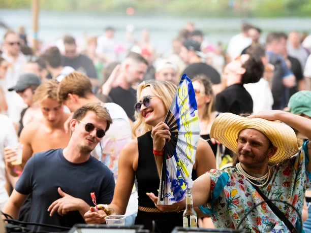 EAT PLAY LOVE Festival Ein fröhliches Festivalpublikum tanzt ausgelassen im Freien. Eine Person in einem farbenfrohen Outfit und großem Strohhut fächert mit einem Fächer Luft zu, während die umgebenden Menschen mit Sonnenbrillen im Sommerlook die Musik genießen. Im Hintergrund verschwimmen blauer Himmel und grüne Bäume.