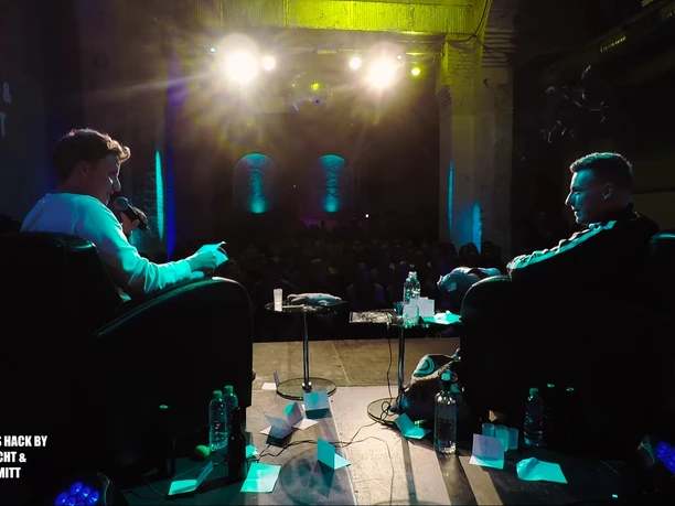 Screenshot1.jpg Photographed from behind: Tomi Schmidt and Felix Lobrecht sit on stage and speak into microphones in the dark background sits the audience