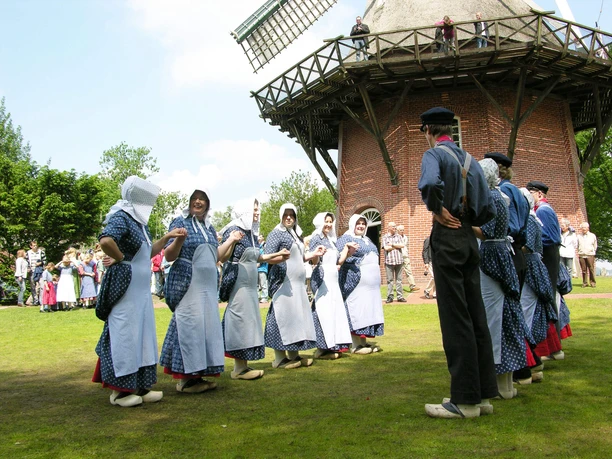 Tag-der-Heimat-Tanzgruppe1.jpg A traditional dance group in blue and white costumes performs a folk dance in front of a historic windmill in Bad Zwischenahn. The dancers wear hoods and aprons while the audience watches in the background.