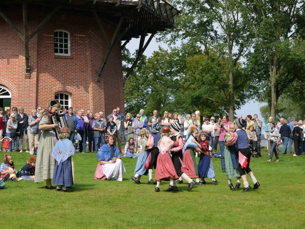 Tag-der-Heimat-Tanzgruppe3.jpg Traditional dance group in colorful costume dancing on a meadow in front of a windmill.