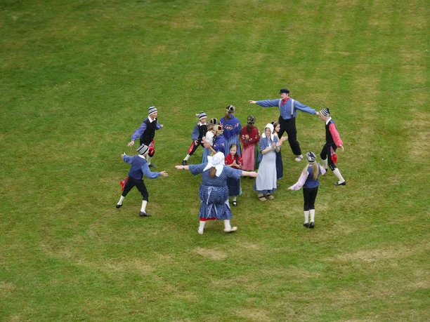 Tag-der-Heimat-Tanzgruppe2.jpg The picture shows a group of traditionally dressed dancers dancing in a circle on a green meadow.