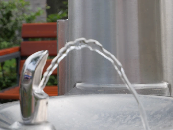 TrinkwasserspenderHwEichenhof 001.JPG Metallener Trinkbrunnen, aus dessen Hahn ein Wasserstrahl in eine runde Schale fließt. Im Hintergrund eine Sitzbank.