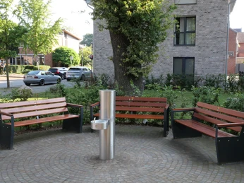 TrinkwasserspenderHwEichenhof 002-1.JPG Drei Holzbänke umgeben einen Trinkbrunnen auf einem gepflasterten Platz, autofreundliche Umgebung.