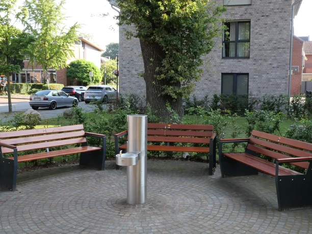 TrinkwasserspenderHwEichenhof 002-1.JPG Drei Holzbänke umgeben einen Trinkbrunnen auf einem gepflasterten Platz, autofreundliche Umgebung.