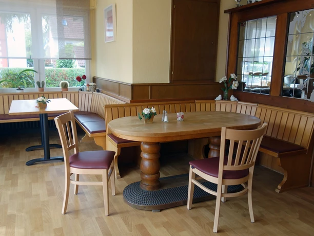 Gaststätte im Hotel Wiedenhof in Hilden Einladender Gastraum mit rundem Holztisch und gepolsterten Stühlen, umgeben von Fenstern und Dekoration.
