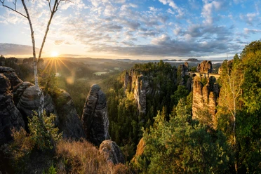 Sonnenaufgang Bastei Felsformationen und eine Brücke in einer grünen Landschaft bei Sonnenaufgang, mit einem klaren Himmel und vereinzelten Wolken.