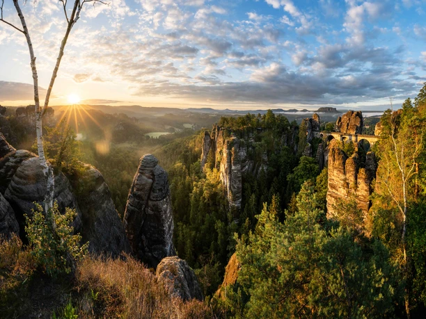 Sonnenaufgang Bastei Felsformationen und eine Brücke in einer grünen Landschaft bei Sonnenaufgang, mit einem klaren Himmel und vereinzelten Wolken.