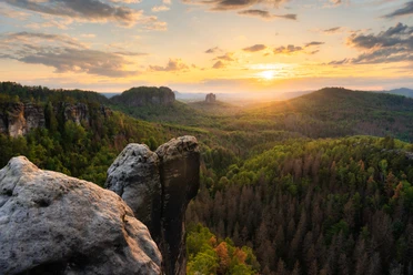 Rock formations in the foreground with a view of green wooded hills at sunset, the sky is partly cloudy and tinted in warm colors.