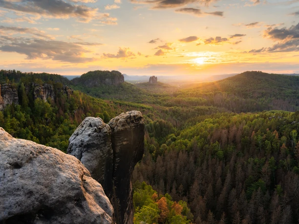 Domerker Felsformationen im Vordergrund mit Blick auf grüne bewaldete Hügel bei Sonnenuntergang, der Himmel ist teils bewölkt und in warmen Farben getönt.