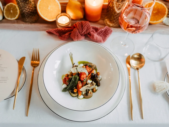Styled Shoot - Rheinloft Cologne Eingedeckter Tisch mit Roséwein im Glas, Gemüse auf einem weißen Teller und orange-goldener DekoTable set with rosé wine in a glass, vegetables on a white plate and orange and gold decorations