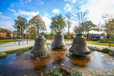 Alter-Kirchplatz_Spelle©MedienAtelierEmsland.jpg Drei gusseiserne Glocken formen einen plätschernden Brunnen auf einem gepflasterten Platz in Spelle.