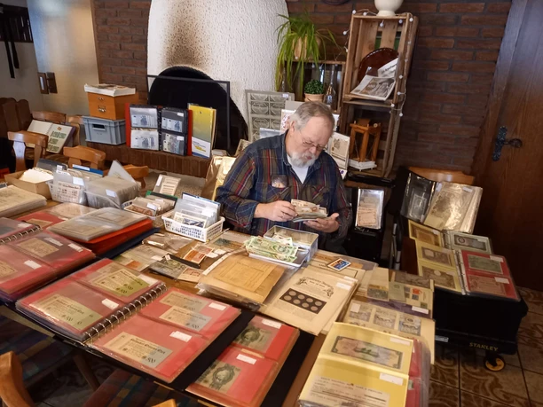 Mann sitzt am Tisch voller Sammelmappen mit historischem Papiergeld in gemütlicher Umgebung.
