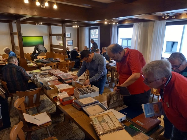 Menschen durchstöbern auf Tischen ausgebreitetes Papiergeld bei einem Sammlertreffen in einem Raum.