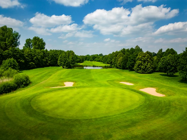 Bahn6.jpg Grüner Golfplatz mit gepflegtem Rasen, umgeben von Bäumen und einem kleinen Teich im Hintergrund.